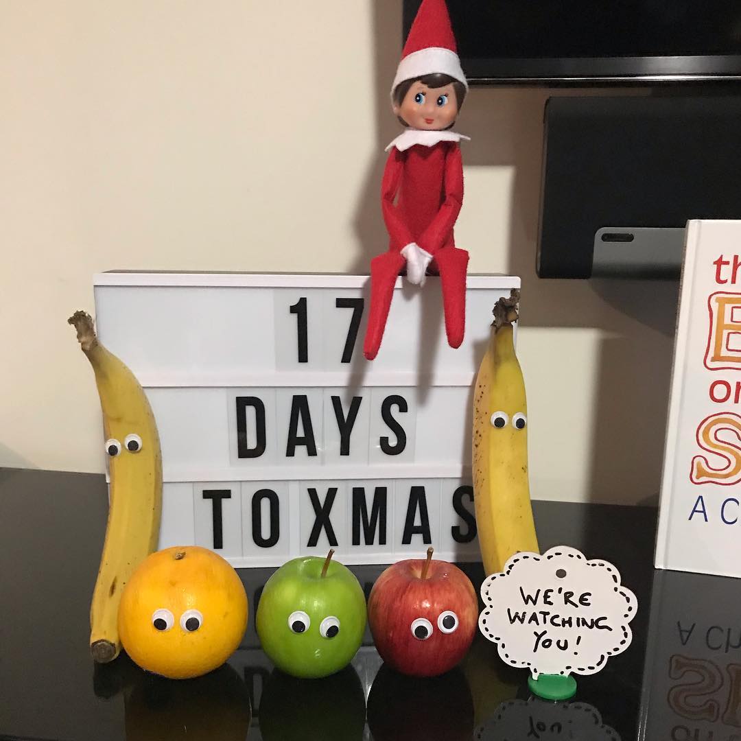 Elf doll sitting on a lightbox reading “17 DAYS TO XMAS,” with googly-eyed bananas and fruit and a sign saying “We’re watching you!”