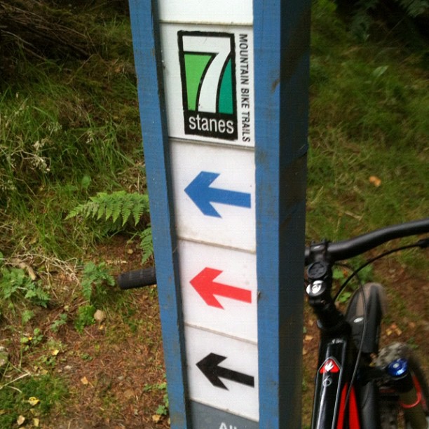 Trail sign marked "7 stanes mountain bike trails" with blue, red, and black left arrows, beside part of a mountain bike.