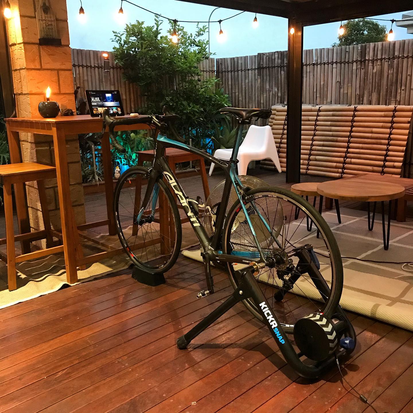 Road bike mounted on an indoor trainer on a covered patio, with string lights, wooden furniture, and a tablet on a table.