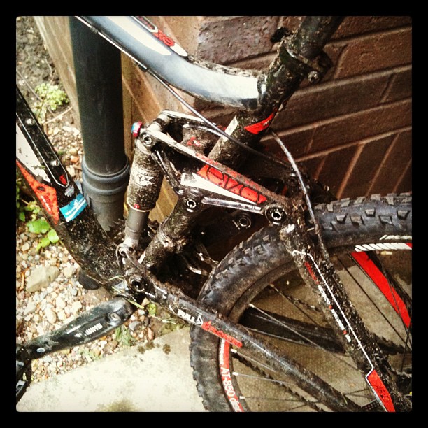 Mud-splattered black mountain bike with rear suspension and thick tires leaning beside a drainpipe and wooden wall.