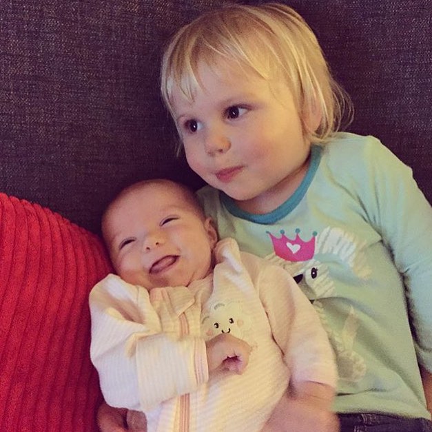 Smiling baby in a light outfit nestled beside an older blond child in a blue shirt on a dark couch.