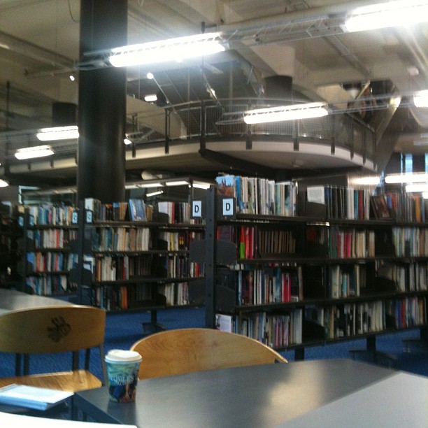 Library interior with bookshelves, tables and chairs, a coffee cup on a table, and an upper-level balcony under bright ceiling lights.