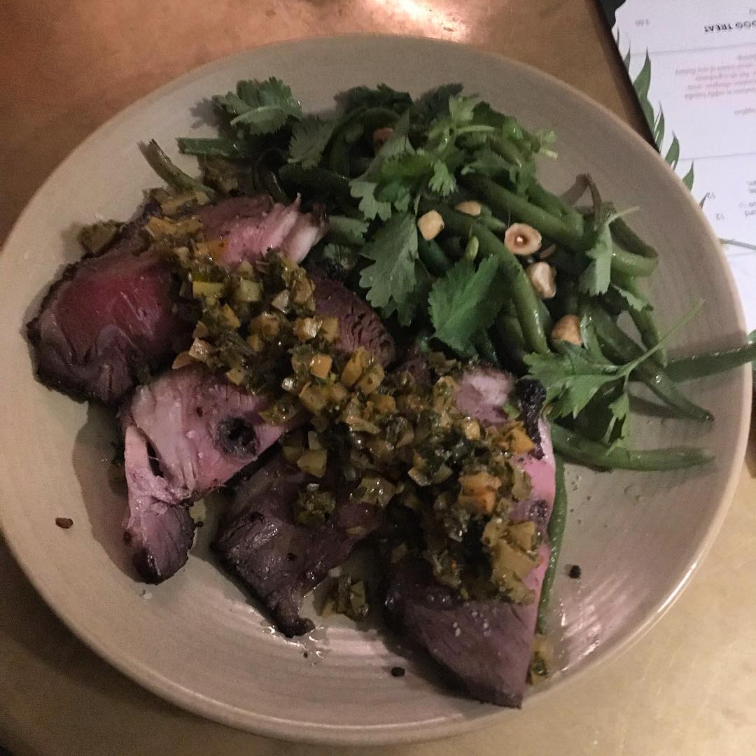 Plate of sliced rare steak topped with herb sauce, served with green beans, cilantro, and chopped nuts.