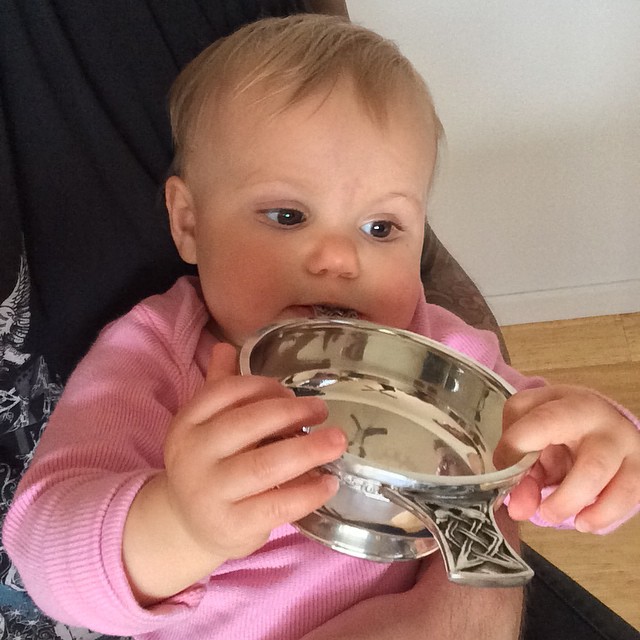 Baby in a pink shirt holding a metal bowl with screws inside and touching the rim with their mouth.