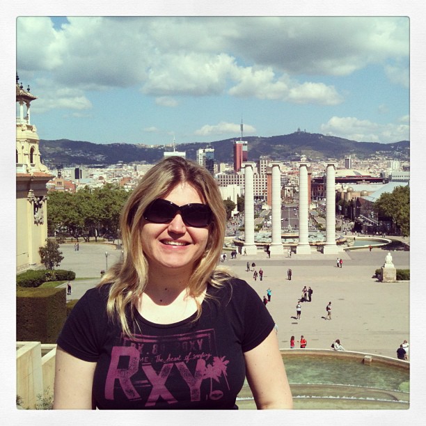 Woman wearing sunglasses and a black T-shirt, smiling in front of a plaza with four white columns and a city skyline.