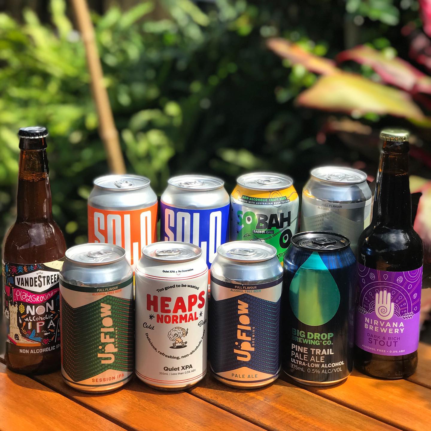 Ten cans and bottles of non-alcoholic and low-alcohol beer arranged on a wooden table outdoors with blurred green plants behind them.