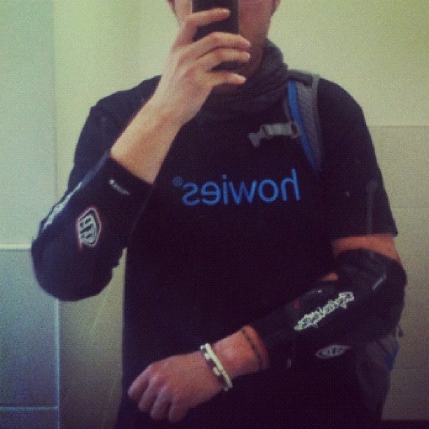 Person taking a mirror selfie, wearing a black shirt, scarf, backpack, and black elbow pads.