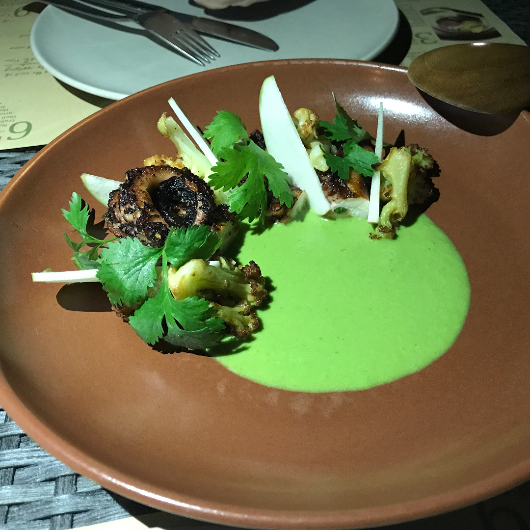 Brown plate with charred cauliflower, white vegetable slices, cilantro, and a pool of bright green sauce on a restaurant table.