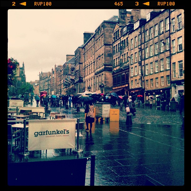 Rainy city street with pedestrians carrying umbrellas, wet pavement, tall stone buildings, and outdoor café barriers in the foreground.