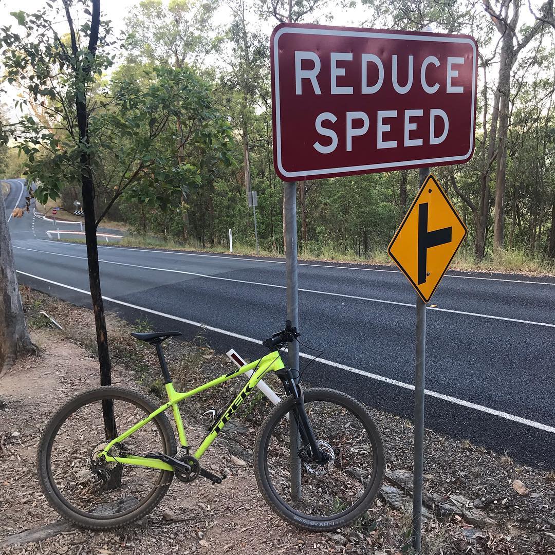 Neon green bicycle beside a roadside Reduce Speed sign and a yellow side-road warning sign, with a paved road and trees in the background.