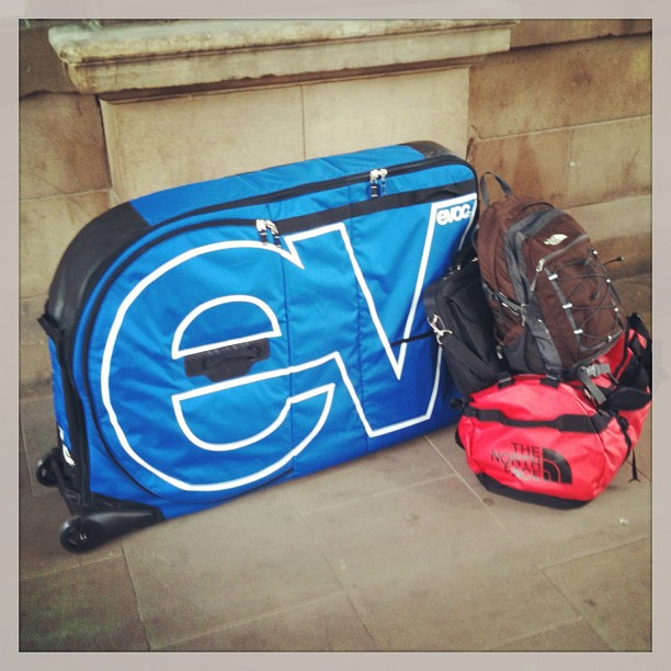 Blue Evoc bike travel case beside a black backpack and red duffel bag on a stone floor against a wall.