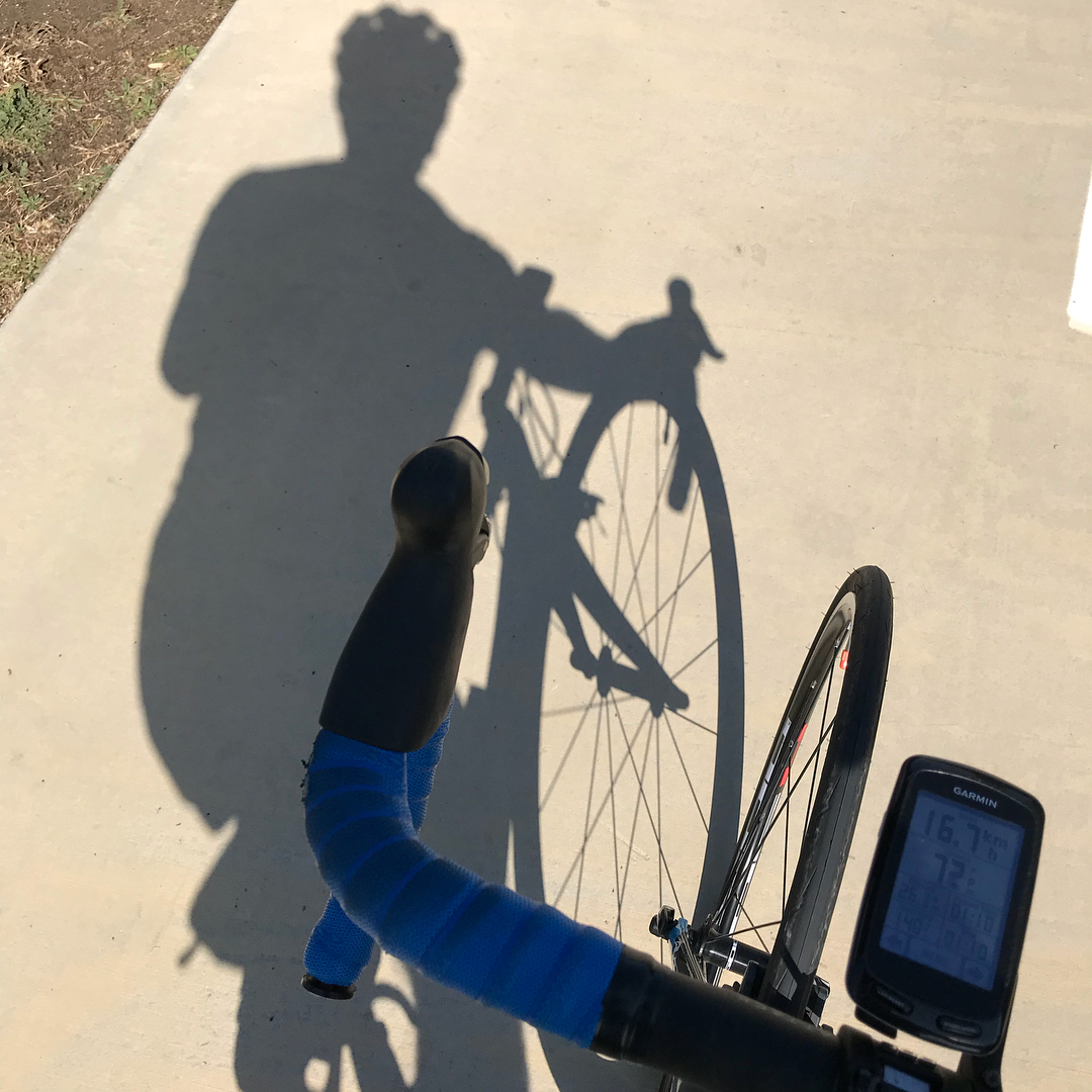 Shadow of a cyclist and bike cast on a sunlit concrete path, with blue handlebar tape, front wheel, and a Garmin display visible.