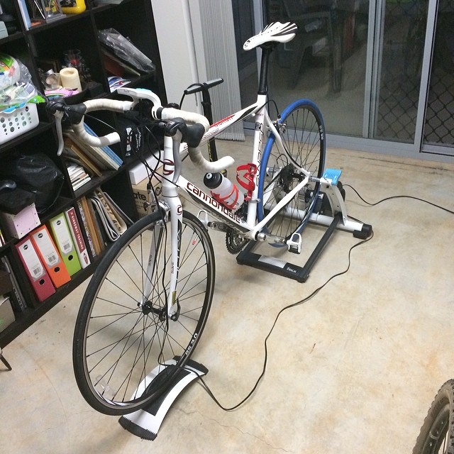 White road bike on an indoor trainer with a front wheel riser, beside a bookshelf and sliding glass door.
