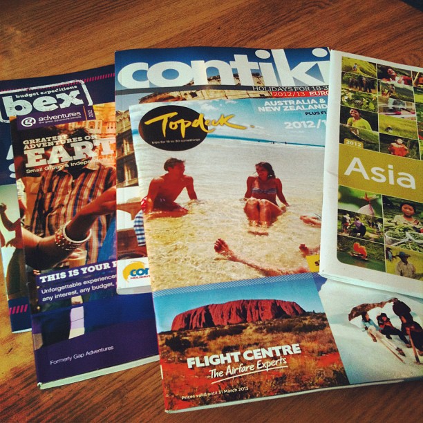 Travel brochures spread on a wooden table, including Contiki, Topdeck, and an Asia guide.