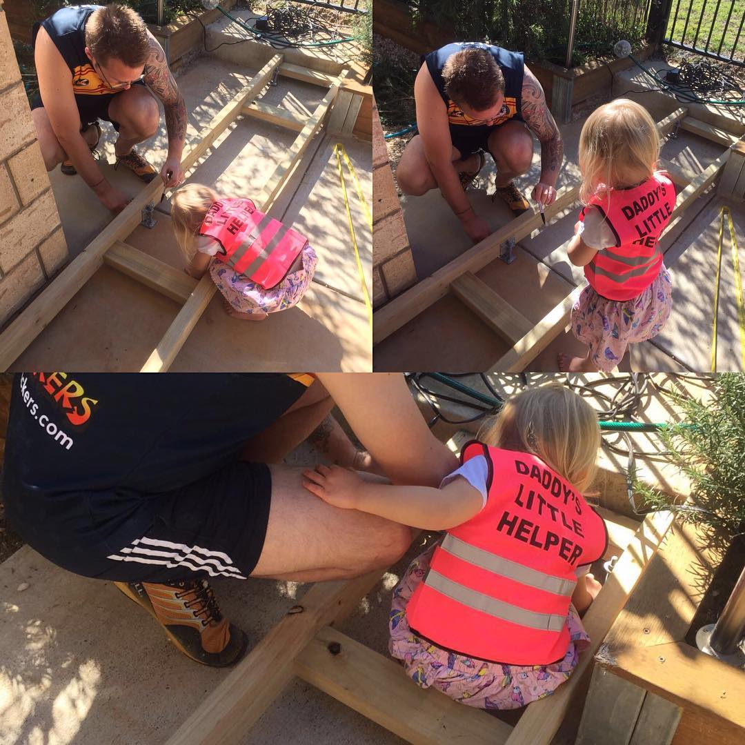 A man and a small child in a bright pink safety vest work together assembling a wooden frame outdoors.