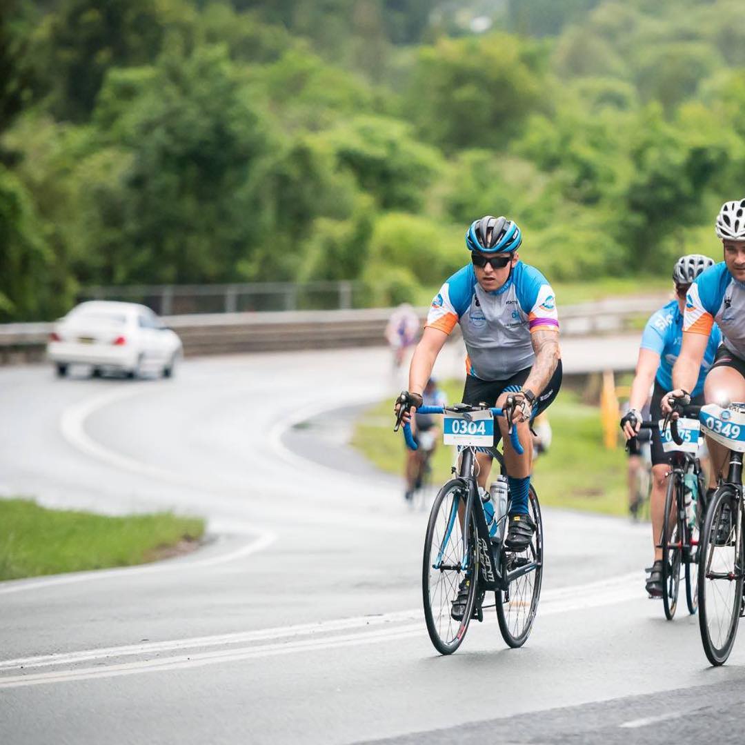 Cyclists wearing helmets ride on a wet, winding road bordered by lush green trees, with a white car in the distance.
