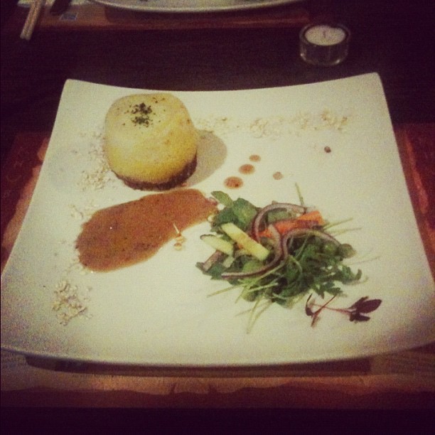 Square white plate with a round savory cake, brown sauce, and a small mixed green salad with sliced onions and vegetables.