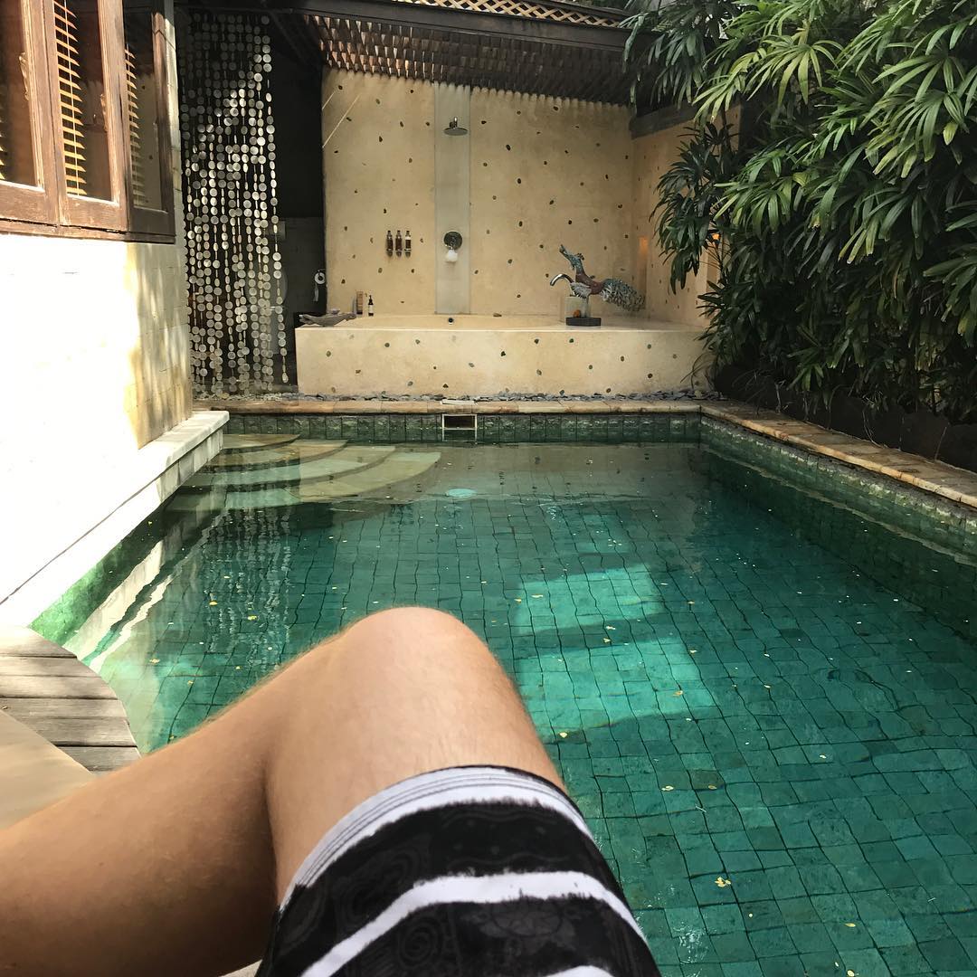 View from poolside with a person’s legs in the foreground, facing a small tiled pool and an outdoor shower beside dense green plants.