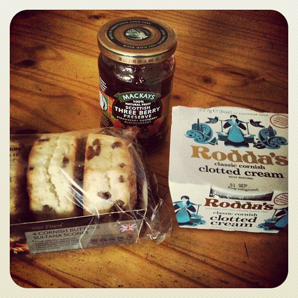 Jar of three berry preserve, packaged sultana scones, and two tubs of clotted cream on a wooden table.