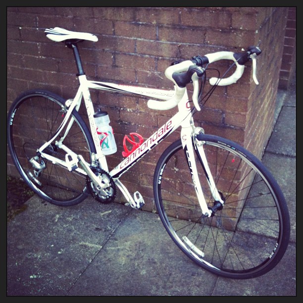 White road bike with drop handlebars, water bottle, and bottle cage leaning against a brick wall on a paved sidewalk.