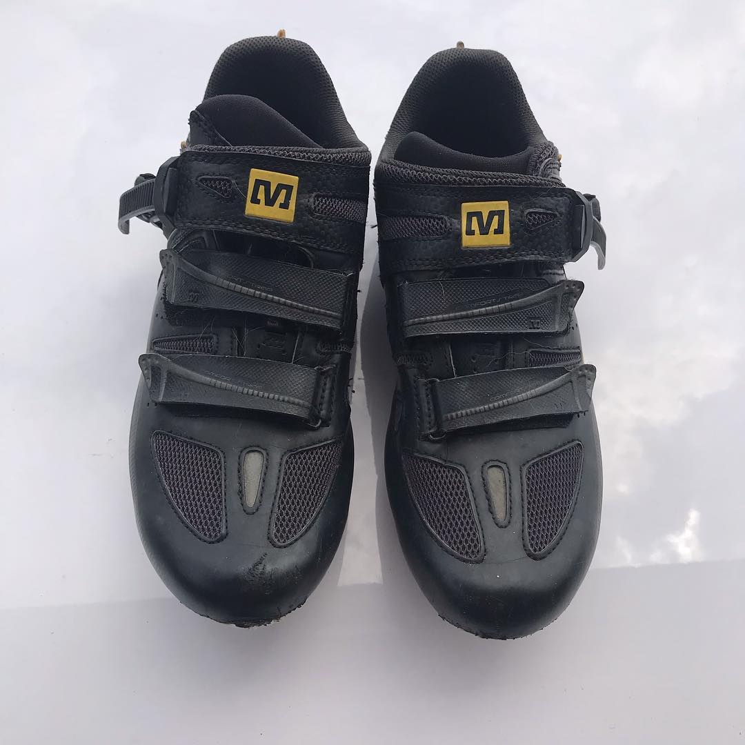 Pair of black cycling shoes with three hook-and-loop straps and yellow logo patches, viewed from above on a white surface.