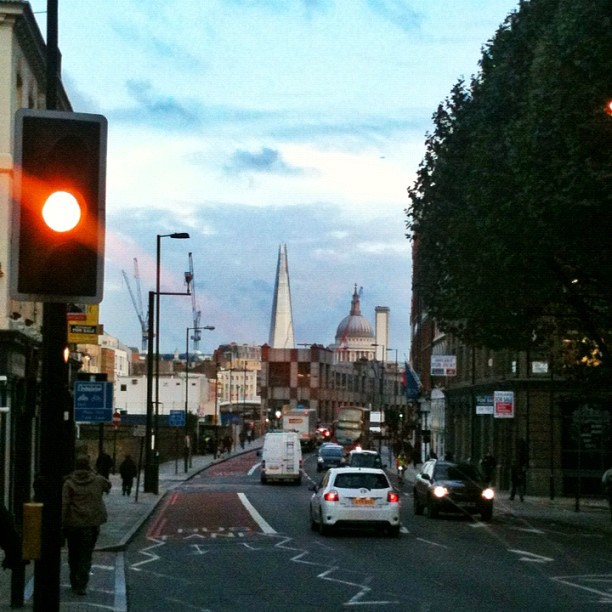 City street at dusk with a red traffic light, cars and a white van, with the Shard and St Paul's Cathedral in the distance.