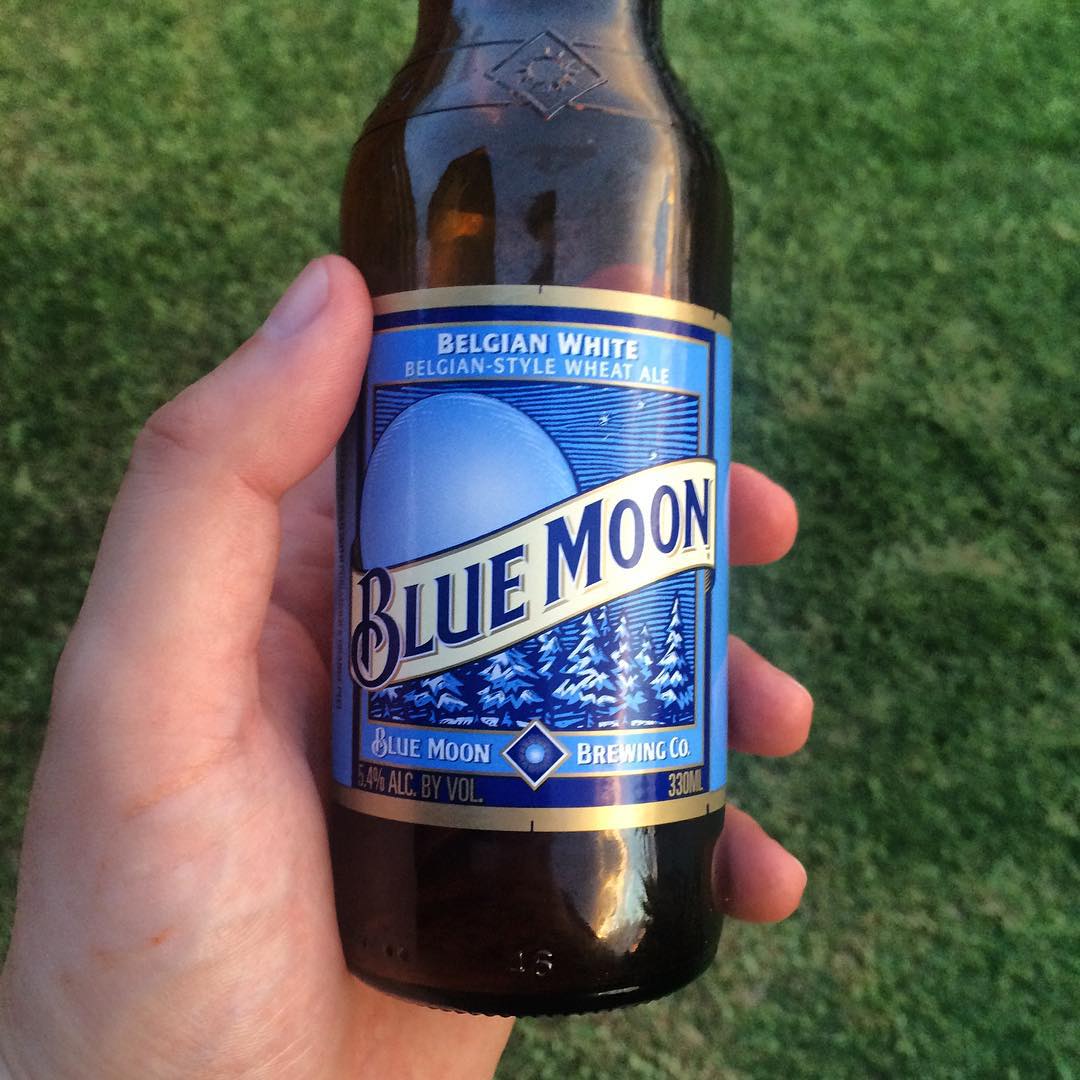 Hand holding a Blue Moon Belgian White beer bottle against a grassy background.