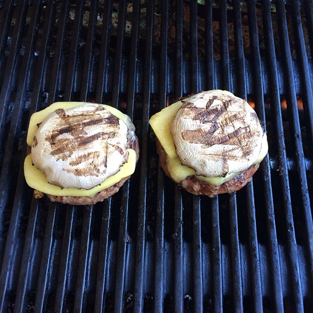 Two burger patties on a grill topped with melted cheese and large grilled mushroom caps.