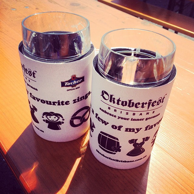 Two plastic cups in white Oktoberfest Brisbane holders sit on a wooden table in bright sunlight.