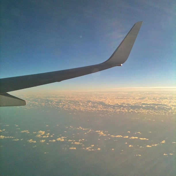 Airplane wing above a blanket of clouds at sunset, with blue sky fading to warm light on the horizon.