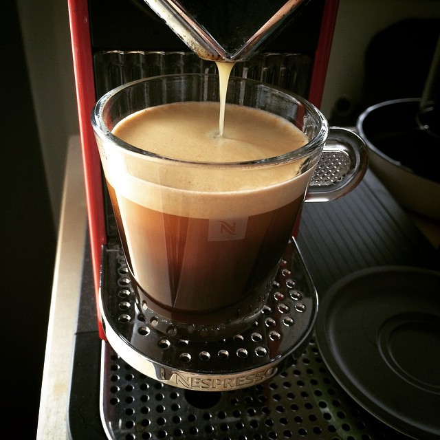 Coffee pouring from a machine into a clear glass cup with thick foam on top.