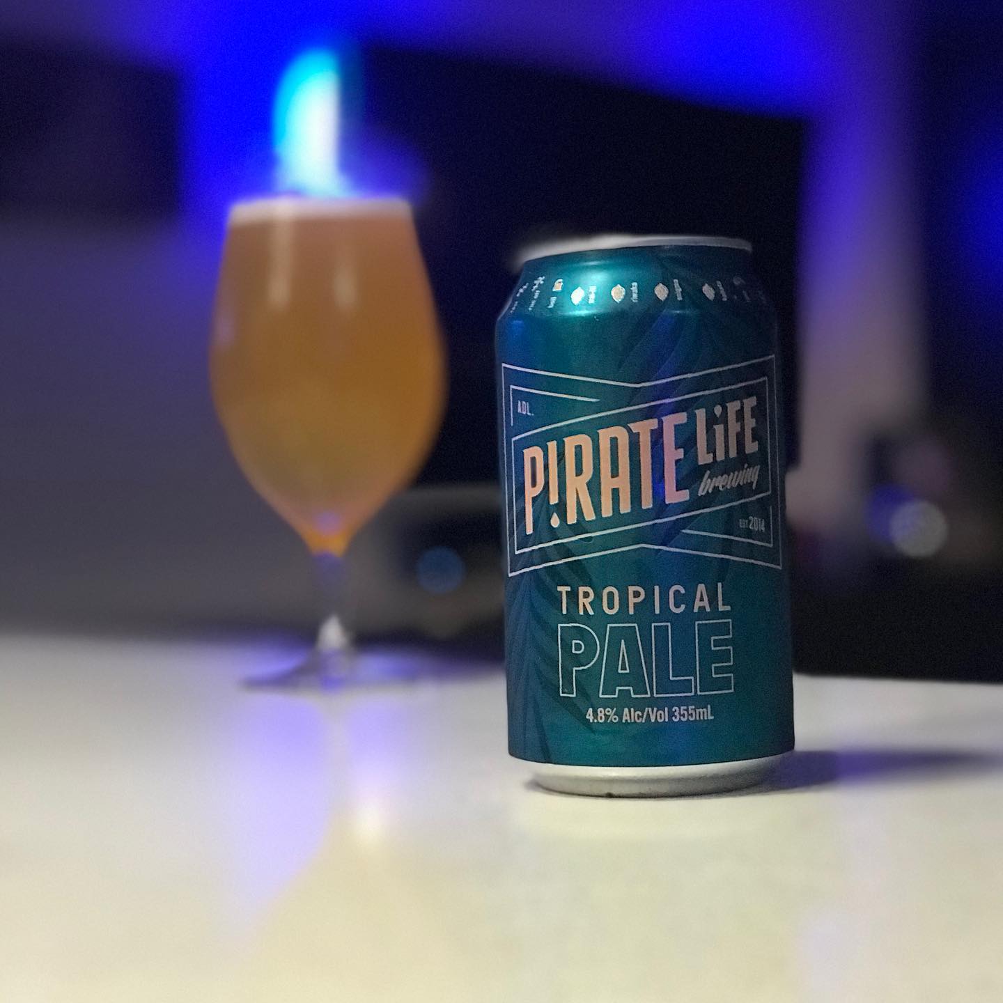 Can of Pirate Life Tropical Pale on a white table, with a blurred glass of hazy beer and blue-purple lights in the background.