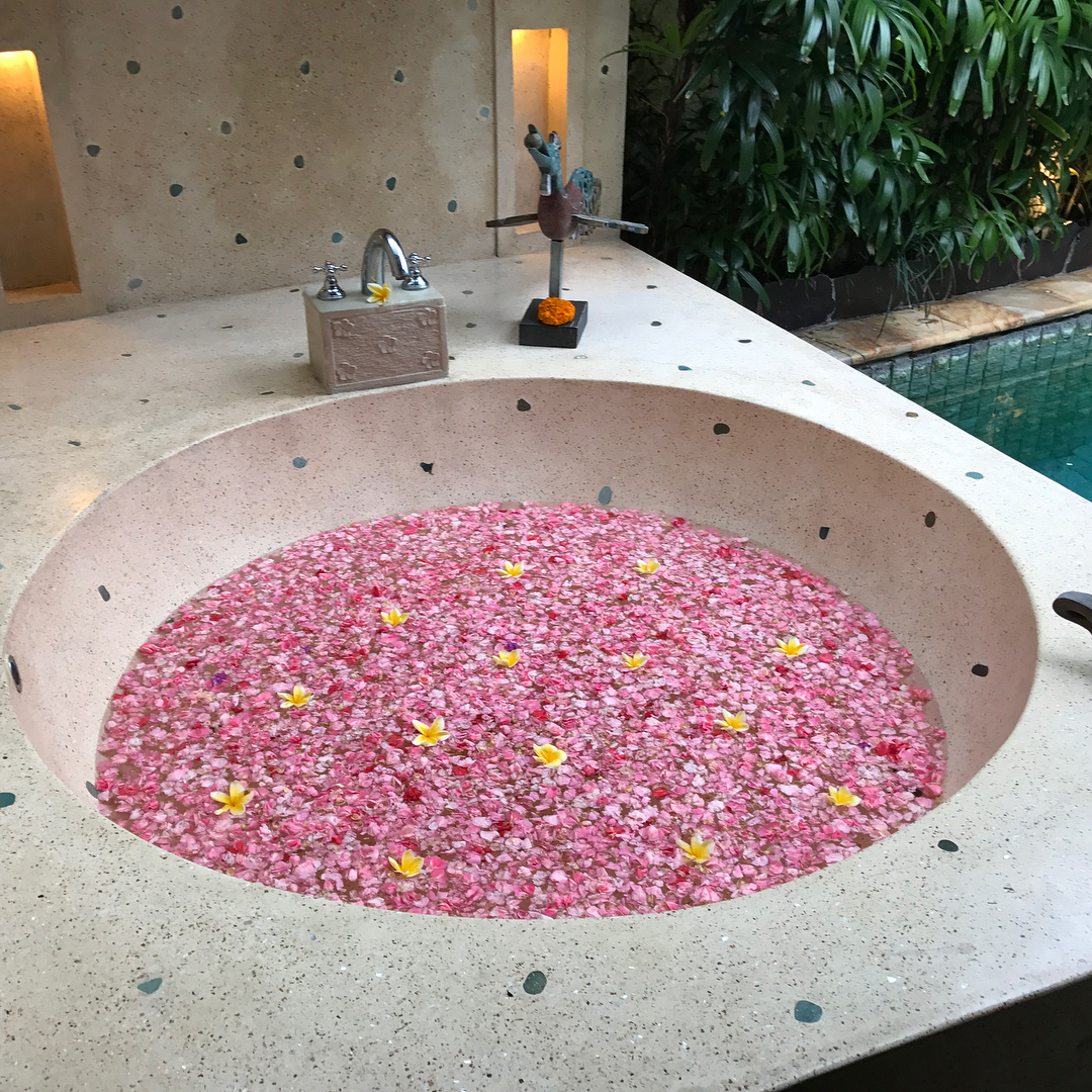 Round stone tub filled with pink flower petals and a few yellow blossoms, beside a faucet, small sculpture, plants, and a pool.