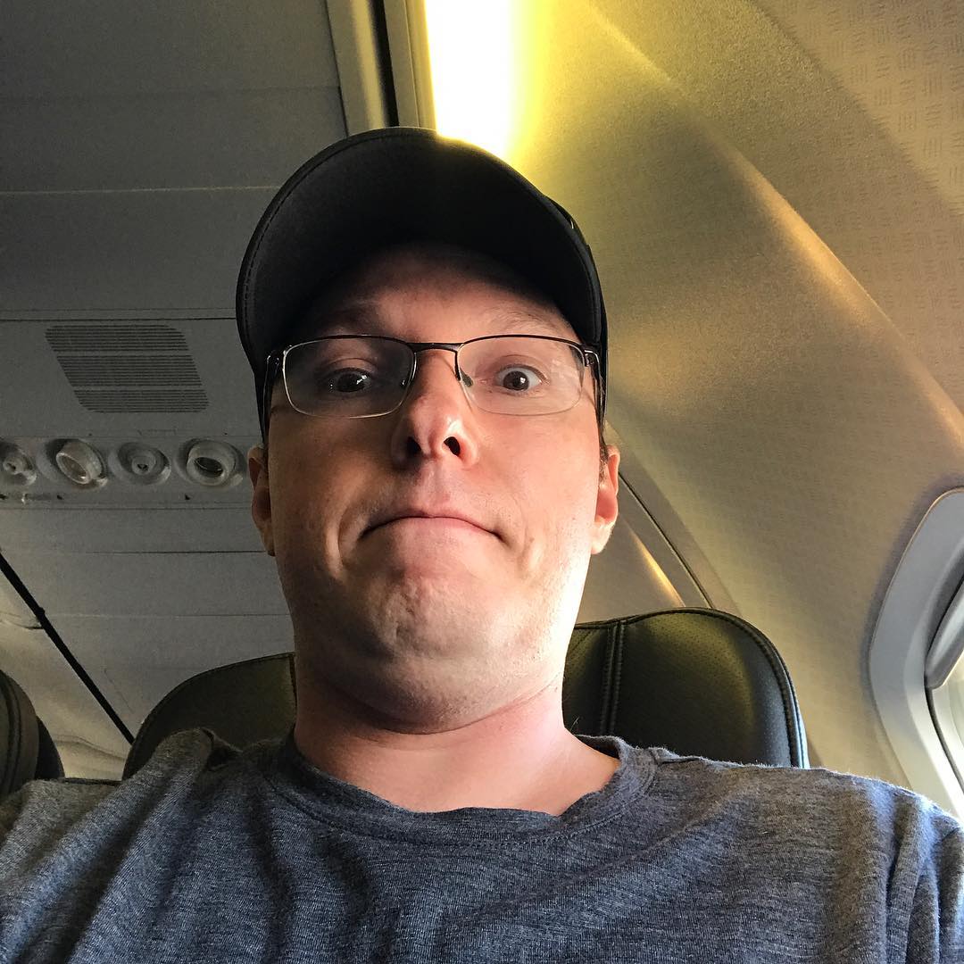 Man wearing glasses and a black cap makes a wide-eyed expression while seated by an airplane window.