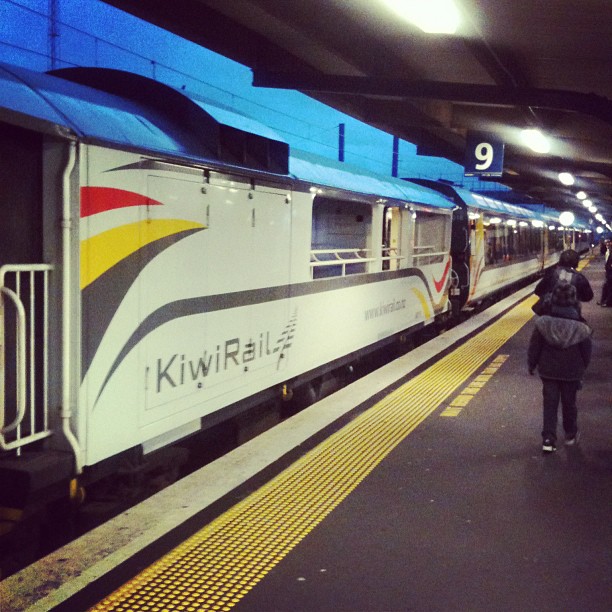KiwiRail train stopped at a covered platform with a few people walking beside it under a sign marked 9.