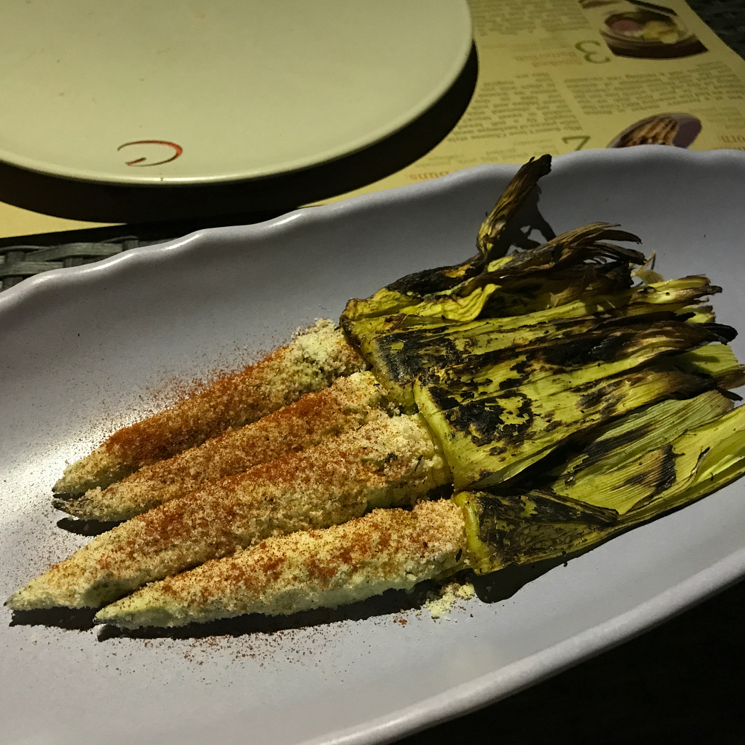 Grilled corn on the cob with charred husks, coated in grated cheese and chili powder on a white platter.
