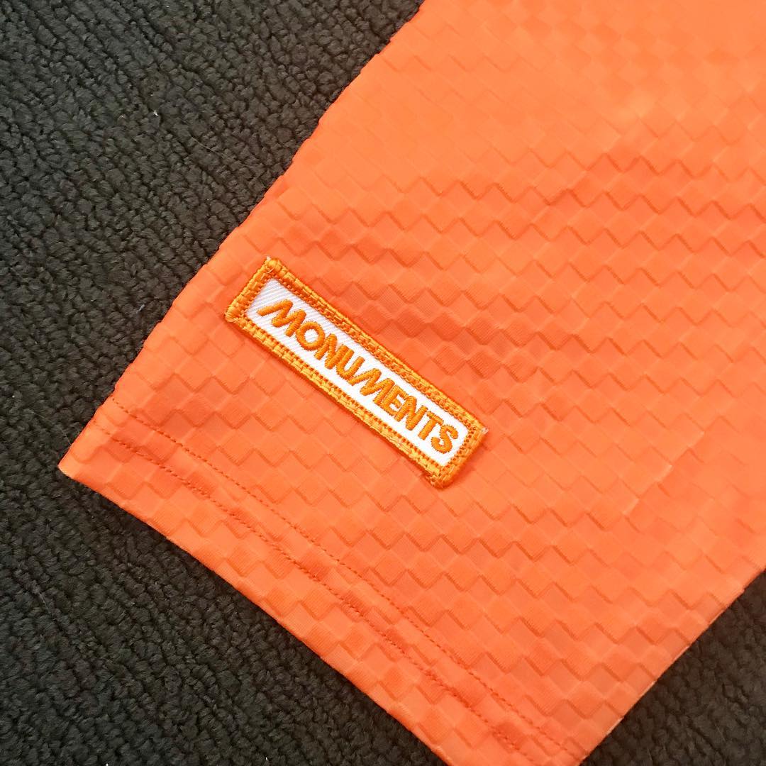 Close-up of orange textured fabric with a stitched white label reading "MONUMENTS" on a dark textured surface.