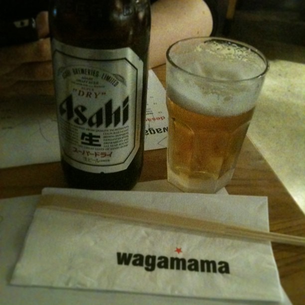 Bottle of Asahi beer, a glass of beer with foam, and chopsticks in a Wagamama napkin on a wooden table.