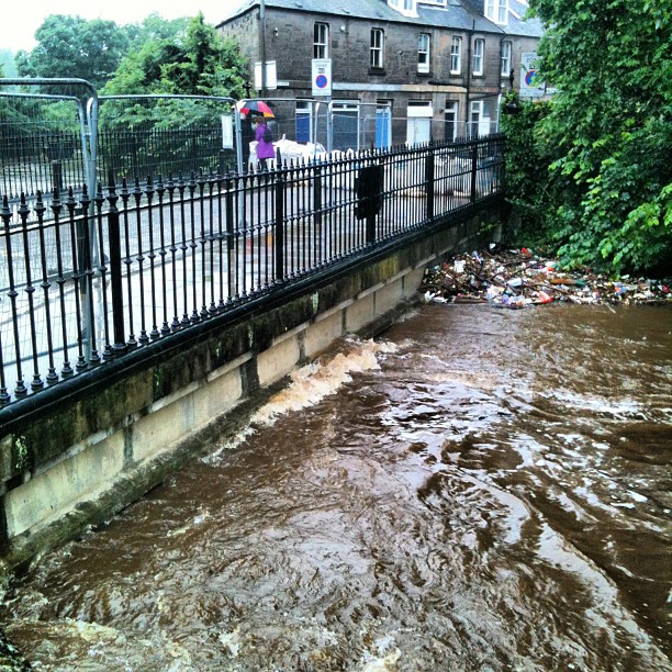 Fast-flowing brown water beside a black iron fence, with debris piled near the bank and a person holding an umbrella on the sidewalk.