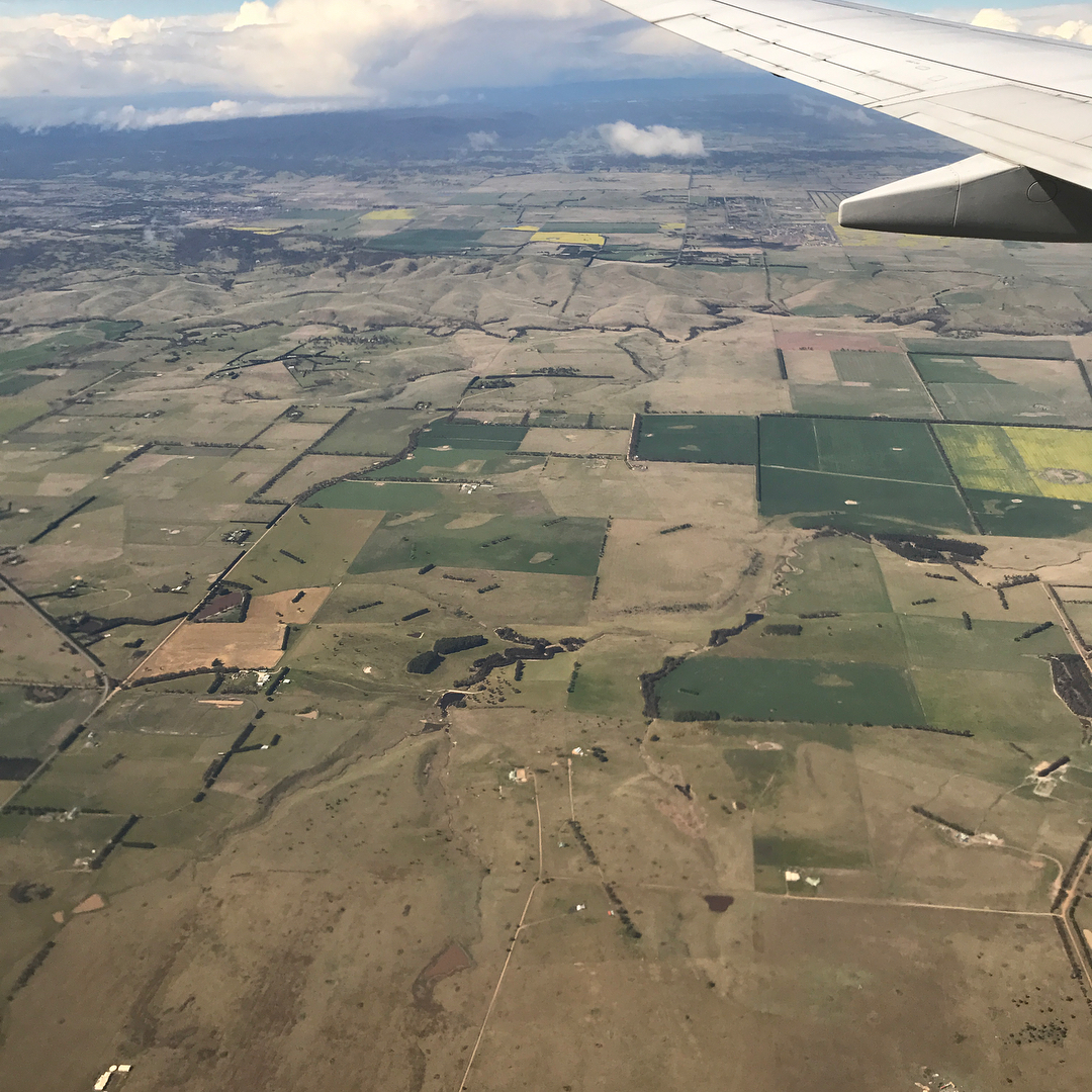 View from an airplane window showing a wing above patchwork fields, rolling hills, and scattered clouds.