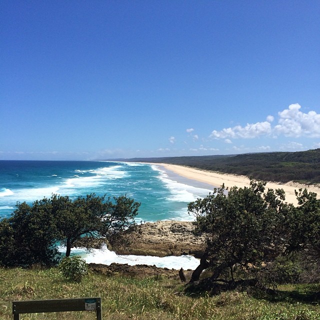 Long sandy beach with turquoise waves, dark green coastal trees, and a clear blue sky with scattered clouds.