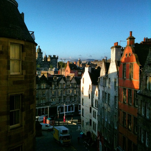 Narrow cobblestone street between tall old buildings, with sunlit facades and a distant city skyline under a clear blue sky.