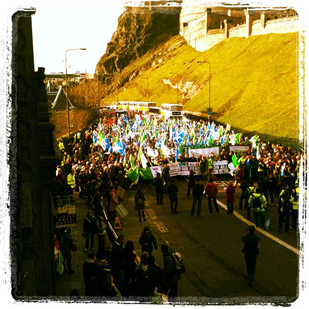 Large street march with a dense crowd carrying blue, green, and white flags and banners between steep grassy and rocky embankments.