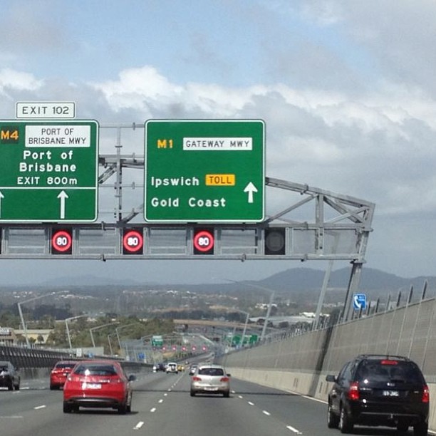 Highway with cars beneath overhead signs for Port of Brisbane, Ipswich, and Gold Coast, with 80 speed limit signals and cloudy sky.