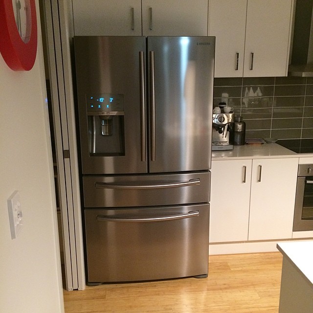 Stainless steel French-door refrigerator with a water dispenser, set between white kitchen cabinets and a countertop coffee machine.