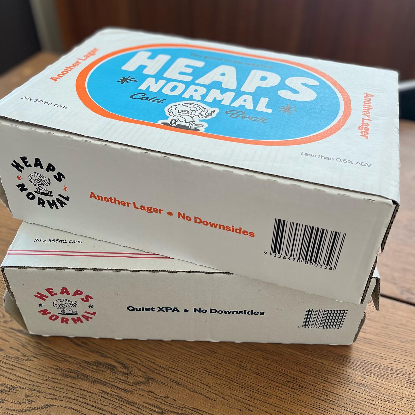 Two stacked Heaps Normal beer cartons on a wooden table, labeled “Another Lager” and “Quiet XPA.”