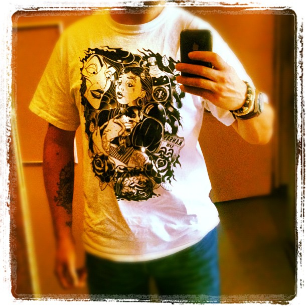 Person taking a mirror selfie in a white graphic T-shirt with black illustrated faces, wearing a watch and showing a forearm tattoo.