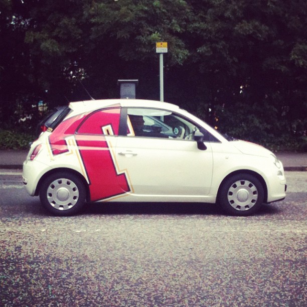 White compact car parked on a street with a large pink and black geometric decal on the side and trees in the background.