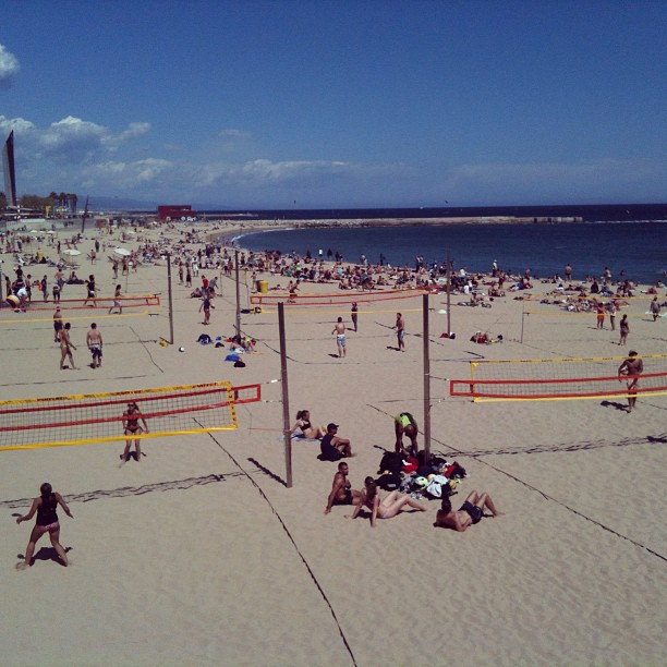 Crowded sandy beach with several volleyball nets, sunbathers, and people swimming along a curved shoreline under a blue sky.