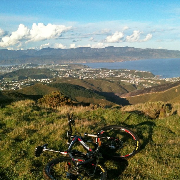 Mountain bike on grassy hillside overlooking rolling hills, a coastal town, and blue water beneath a cloudy sky.
